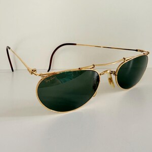 K&ouml;nnte beinhalten: Goldfarbene Sonnenbrille mit dunkelgr&uuml;nen Gl&auml;sern. Die Sonnenbrille hat ein einzigartiges Design mit einem gebogenen oberen B&uuml;gel und einem Ray-Ban-Logo auf dem Glas. Die B&uuml;gel sind d&uuml;nn und haben eine braune Spitze. Auf wei&szlig;em Hintergrund.