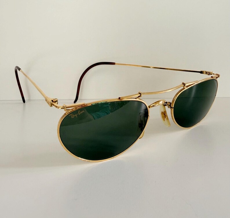 K&ouml;nnte beinhalten: Goldfarbene Sonnenbrille mit dunkelgr&uuml;nen Gl&auml;sern. Die Sonnenbrille hat ein einzigartiges Design mit einem gebogenen oberen B&uuml;gel und einem Ray-Ban-Logo auf dem Glas. Die B&uuml;gel sind d&uuml;nn und haben eine braune Spitze. Auf wei&szlig;em Hintergrund.