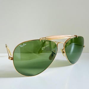 May include: Gold-framed Ray-Ban sunglasses with green lenses. The sunglasses have a bridge bar and a light-colored brow bar. The Ray-Ban logo is visible on the left lens. The sunglasses are on a white surface.