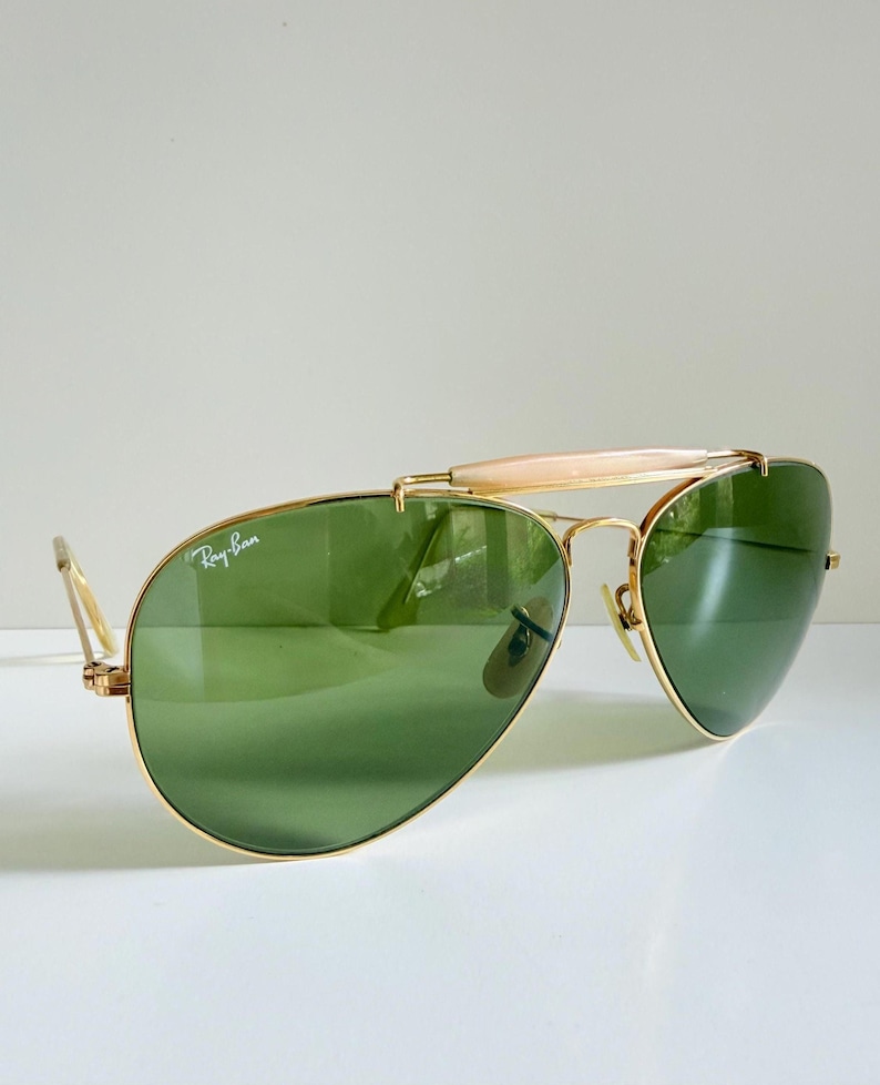 May include: Gold-framed Ray-Ban sunglasses with green lenses. The sunglasses have a bridge bar and a light-colored brow bar. The Ray-Ban logo is visible on the left lens. The sunglasses are on a white surface.