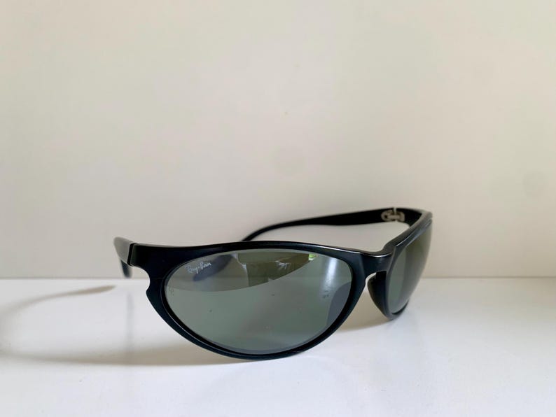 May include: Black sunglasses featuring a wrap-around frame design with dark tinted lenses. The Ray-Ban logo is printed in white on the upper corner of the right lens. The frame has a smooth, matte finish with curved temples and integrated nose pads.
