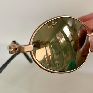 May include: Gold-tone Ray-Ban sunglasses with oval lenses. The lenses reflect a room with a window. The frames are gold with black arms. The Ray-Ban logo is visible on the lens.