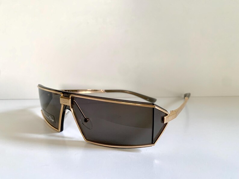 Peut inclure: Lunettes de soleil avec des verres fonc&eacute;s et une monture dor&eacute;e. Les lunettes ont une forme g&eacute;om&eacute;trique unique avec un dessus droit et un bas inclin&eacute;. Les branches sont fines et dor&eacute;es, avec une courbe subtile.