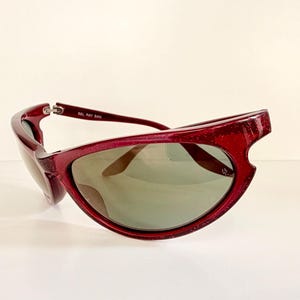 May include: A pair of burgundy sunglasses with a unique, angular frame design. The lenses are a muted green, and the text "BAL RAY BAN" is visible on the temple. These sunglasses offer a stylish, retro aesthetic.