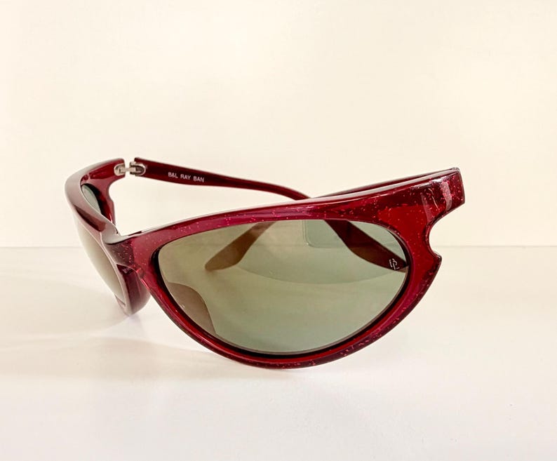 May include: A pair of burgundy sunglasses with a unique, angular frame design. The lenses are a muted green, and the text "BAL RAY BAN" is visible on the temple. These sunglasses offer a stylish, retro aesthetic.