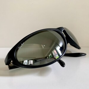 May include: Black sunglasses with a matte finish and dark, reflective lenses. The left lens features the white Ray-Ban logo printed in the upper corner. The frames have an oval shape with thick temples, and the design is captured from a low angle.