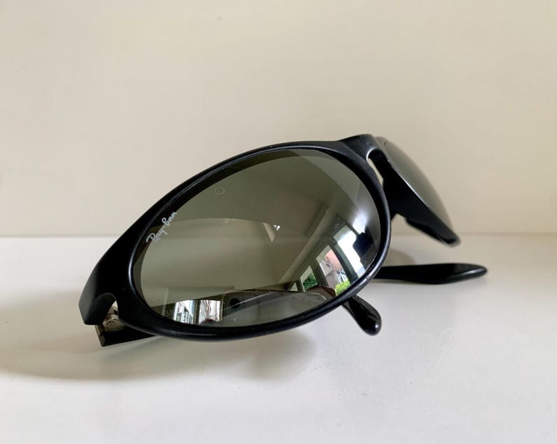 May include: Black sunglasses with a matte finish and dark, reflective lenses. The left lens features the white Ray-Ban logo printed in the upper corner. The frames have an oval shape with thick temples, and the design is captured from a low angle.