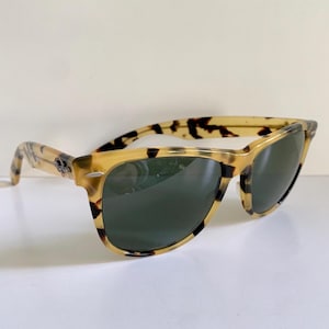 May include: Tortoiseshell sunglasses with dark green lenses. The frames have a classic design with a light brown and black pattern. The sunglasses are set against a white background.