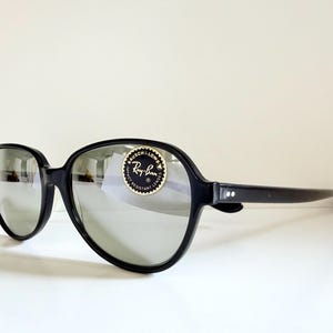 May include: Black sunglasses with large, rounded frames and reflective lenses. The sunglasses have a Ray-Ban sticker on the lens. The arms of the sunglasses are straight and have two small white dots near the hinge.