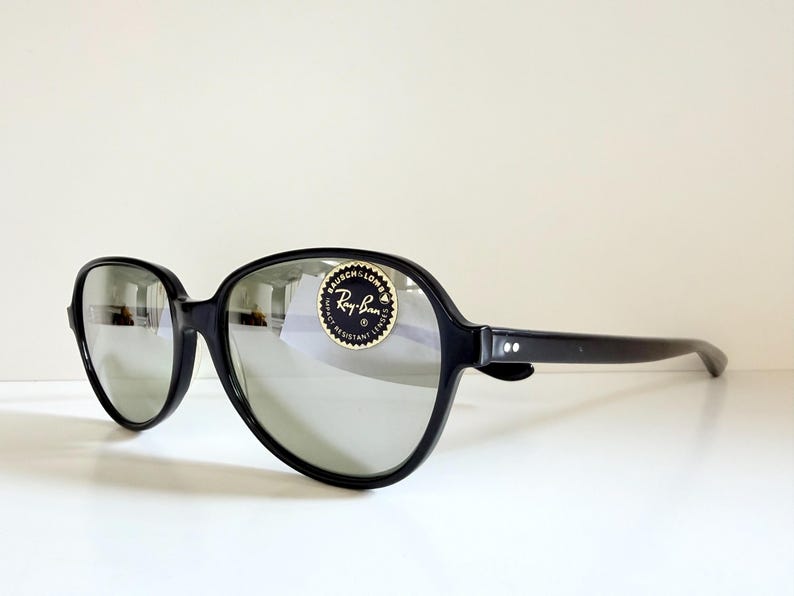 May include: Black sunglasses with large, rounded frames and reflective lenses. The sunglasses have a Ray-Ban sticker on the lens. The arms of the sunglasses are straight and have two small white dots near the hinge.