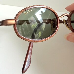 May include: Brown oval-framed sunglasses with green lenses. The Ray-Ban logo is visible on the lens. The frame has a marbled effect. The sunglasses have a curved bridge and temples.