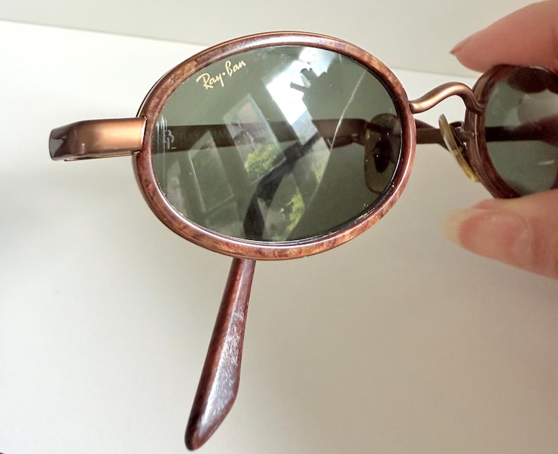 May include: Brown oval-framed sunglasses with green lenses. The Ray-Ban logo is visible on the lens. The frame has a marbled effect. The sunglasses have a curved bridge and temples.