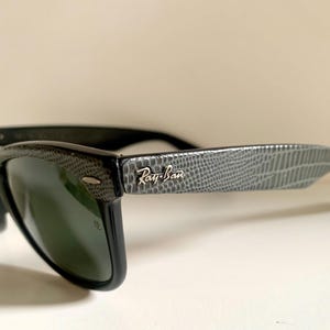 May include: Black Ray-Ban sunglasses with a textured gray pattern on the temples. The Ray-Ban logo is visible on the temple. The lenses are dark green.