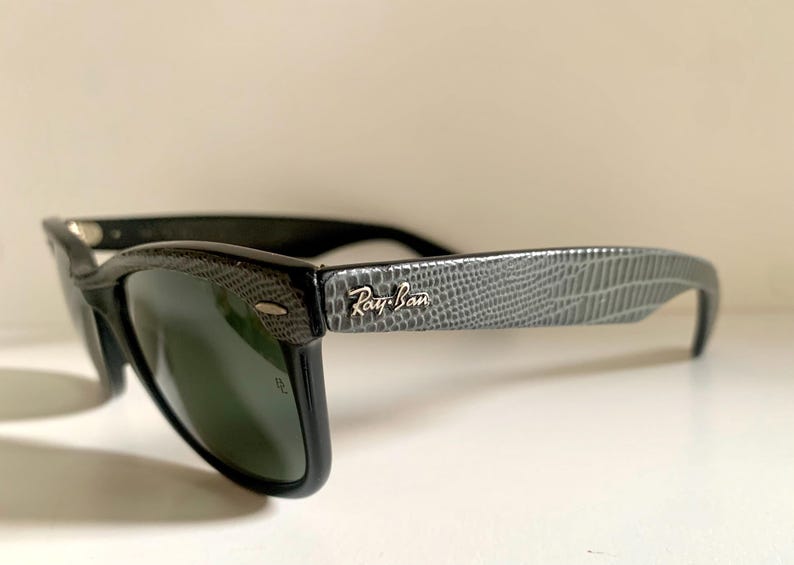 May include: Black Ray-Ban sunglasses with a textured gray pattern on the temples. The Ray-Ban logo is visible on the temple. The lenses are dark green.