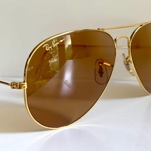 May include: Gold-coloured aviator sunglasses with brown lenses. The Ray-Ban logo is visible on the upper left lens. The sunglasses have a double bridge and a classic teardrop shape, reflecting light.