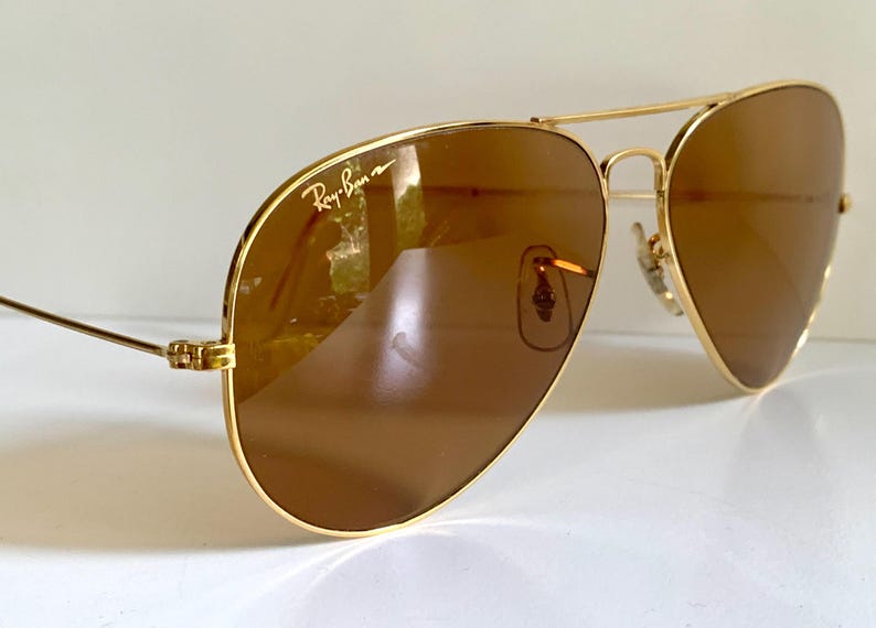 May include: Gold-coloured aviator sunglasses with brown lenses. The Ray-Ban logo is visible on the upper left lens. The sunglasses have a double bridge and a classic teardrop shape, reflecting light.