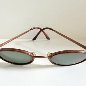 May include: Vintage oval sunglasses with a bronze metal frame and green-tinted lenses. The arms are thin and feature the text "B&L RAY BAN". A classic accessory for eye protection.