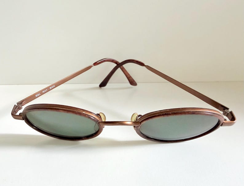 May include: Vintage oval sunglasses with a bronze metal frame and green-tinted lenses. The arms are thin and feature the text "B&L RAY BAN". A classic accessory for eye protection.