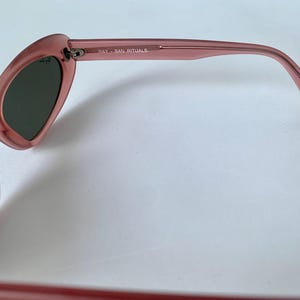 Peut inclure: Gros plan sur des lunettes de soleil roses avec des verres fonc&eacute;s. La monture est en plastique rose translucide. Le texte "RAY-BAN RITUALS" est imprim&eacute; sur la branche. Les lunettes de soleil sont pos&eacute;es sur un fond blanc.