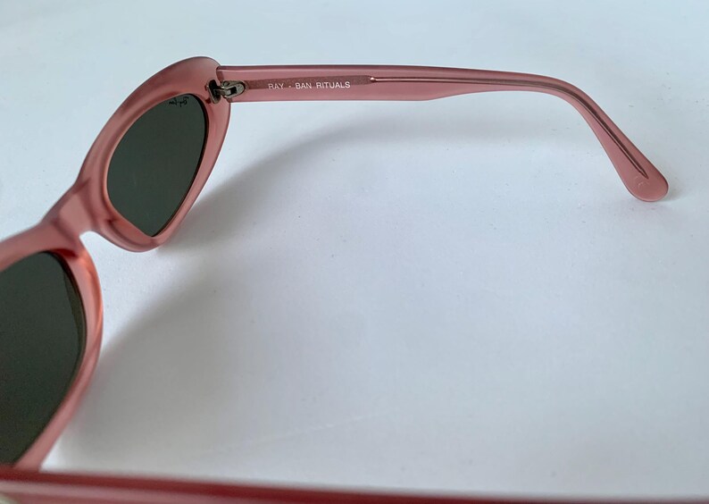 Peut inclure: Gros plan sur des lunettes de soleil roses avec des verres fonc&eacute;s. La monture est en plastique rose translucide. Le texte "RAY-BAN RITUALS" est imprim&eacute; sur la branche. Les lunettes de soleil sont pos&eacute;es sur un fond blanc.