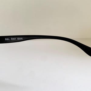May include: A black eyeglass temple arm featuring a silver-toned metal hinge and the white printed text B&L RAY BAN on the inner side. The curved plastic arm tapers toward the end, which displays a small embossed CE mark near the tip.