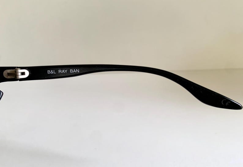 May include: A black eyeglass temple arm featuring a silver-toned metal hinge and the white printed text B&L RAY BAN on the inner side. The curved plastic arm tapers toward the end, which displays a small embossed CE mark near the tip.