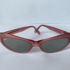 Peut inclure: Lunettes de soleil &agrave; monture rose avec verres gris fonc&eacute;. Les lunettes ont une forme ovale incurv&eacute;e et un logo Ray-Ban sur la lentille gauche. Les montures sont de couleur rose clair, et les branches se rejoignent au niveau du pont.