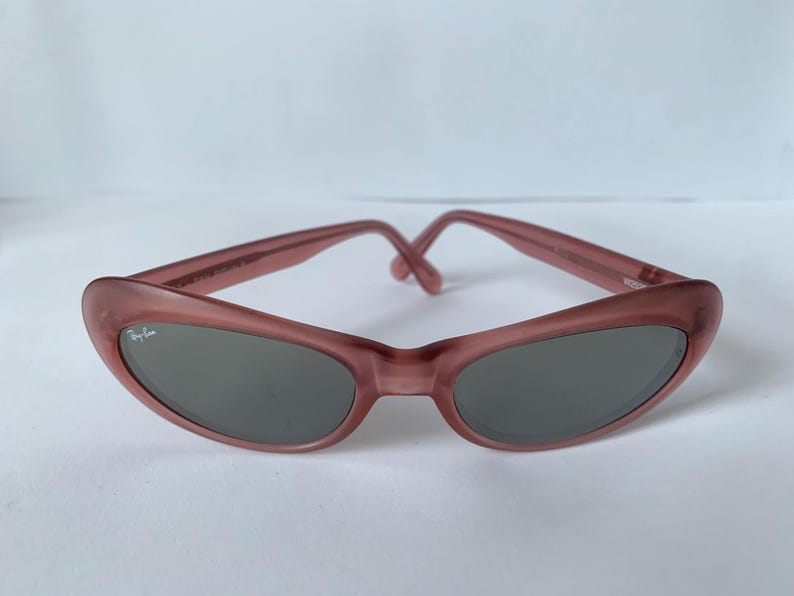 Peut inclure: Lunettes de soleil &agrave; monture rose avec verres gris fonc&eacute;. Les lunettes ont une forme ovale incurv&eacute;e et un logo Ray-Ban sur la lentille gauche. Les montures sont de couleur rose clair, et les branches se rejoignent au niveau du pont.