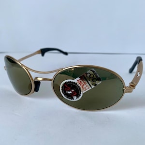 May include: Gold-framed sunglasses with olive-green lenses. The sunglasses have a sporty design with black accents on the temples and nose bridge. A sticker with the word "SPORT" is visible on one lens.