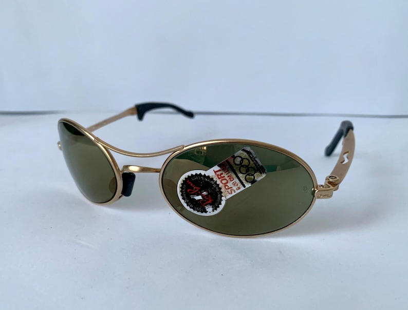 May include: Gold-framed sunglasses with olive-green lenses. The sunglasses have a sporty design with black accents on the temples and nose bridge. A sticker with the word "SPORT" is visible on one lens.