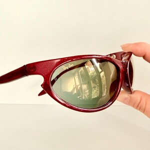 May include: Maroon-colored Ray-Ban sunglasses with a curved frame and green-tinted lenses. The Ray-Ban logo is visible on the lens. The sunglasses are held by a hand.