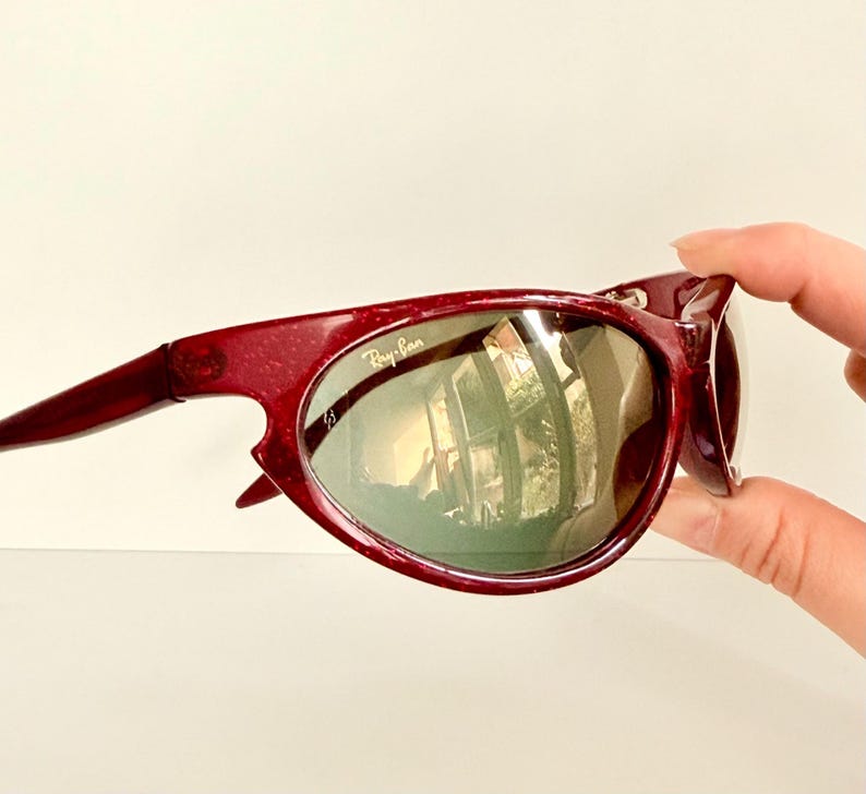 May include: Maroon-colored Ray-Ban sunglasses with a curved frame and green-tinted lenses. The Ray-Ban logo is visible on the lens. The sunglasses are held by a hand.