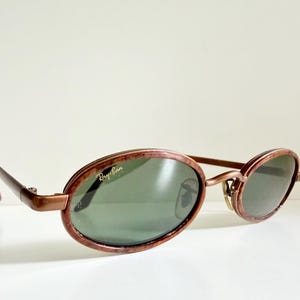 May include: Oval sunglasses with a brown frame and green lenses. The Ray-Ban logo is visible on the lens. The sunglasses have a retro style.