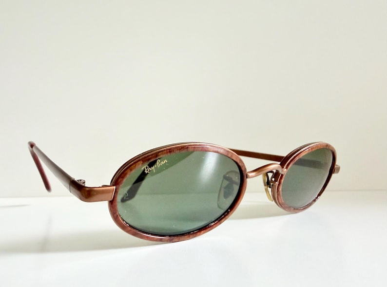 May include: Oval sunglasses with a brown frame and green lenses. The Ray-Ban logo is visible on the lens. The sunglasses have a retro style.