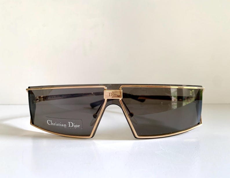 Peut inclure: Lunettes de soleil rectangulaires avec une monture dor&eacute;e et des verres fonc&eacute;s. Le texte "Christian Dior" est visible sur les verres. La monture pr&eacute;sente un accent dor&eacute; et un petit logo "Dior".