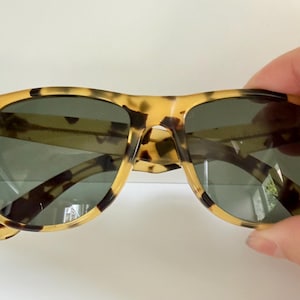 May include: Tortoiseshell sunglasses with dark lenses. The frames have a classic, rectangular shape with a speckled pattern in shades of tan and brown. The sunglasses are held by a hand.