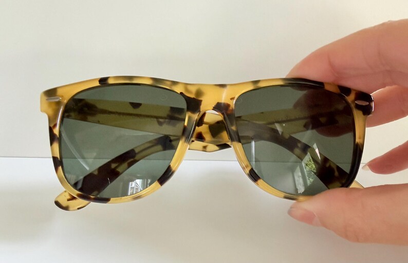 May include: Tortoiseshell sunglasses with dark lenses. The frames have a classic, rectangular shape with a speckled pattern in shades of tan and brown. The sunglasses are held by a hand.