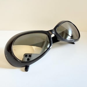 May include: Black sunglasses with a rounded, oversized frame. The lenses are reflective, and the sunglasses have a sleek, modern design. The sunglasses are set against a neutral background.
