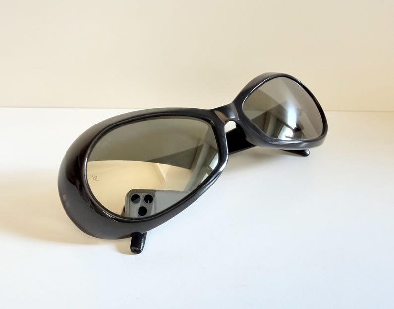 May include: Black sunglasses with a rounded, oversized frame. The lenses are reflective, and the sunglasses have a sleek, modern design. The sunglasses are set against a neutral background.