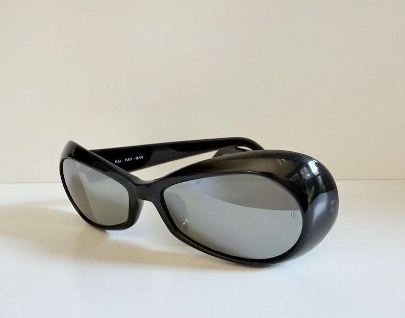 May include: Black sunglasses with a curved, wraparound design. The lenses are reflective silver, and the frame is glossy black. The text "B&L RAY-BAN" is visible on the frame. The sunglasses are set against a plain white background.