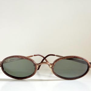 May include: Oval sunglasses with a brown tortoiseshell frame and dark green lenses. The sunglasses feature a thin, gold-coloured bridge and temples. The lenses are oval, and the frame has a subtle, textured pattern.