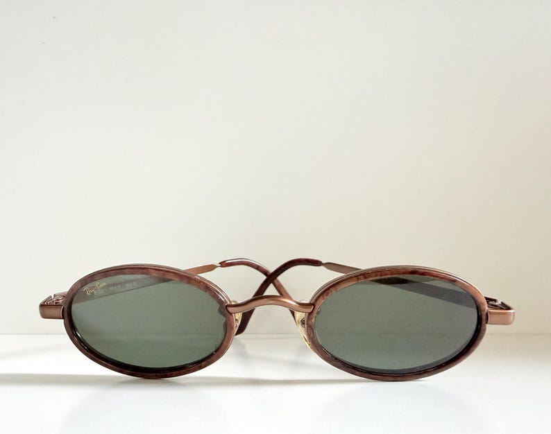 May include: Oval sunglasses with a brown tortoiseshell frame and dark green lenses. The sunglasses feature a thin, gold-coloured bridge and temples. The lenses are oval, and the frame has a subtle, textured pattern.