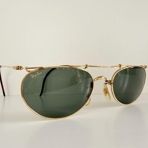 K&ouml;nnte beinhalten: Goldfarbene Sonnenbrille mit dunkelgr&uuml;nen Gl&auml;sern. Die Sonnenbrille hat ein einzigartiges Design mit einem Steg, der &uuml;ber den Gl&auml;sern liegt. Das Ray-Ban-Logo ist sichtbar. Die B&uuml;gel sind d&uuml;nn und goldfarben.