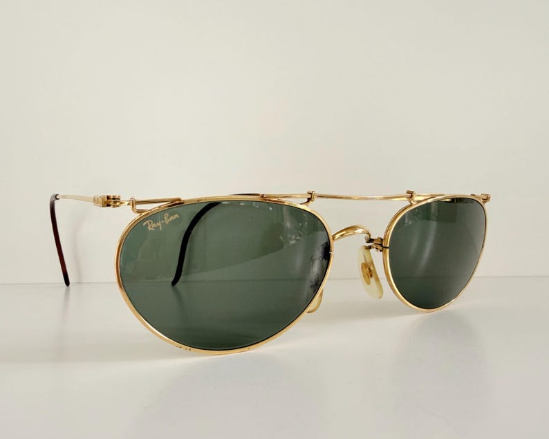 K&ouml;nnte beinhalten: Goldfarbene Sonnenbrille mit dunkelgr&uuml;nen Gl&auml;sern. Die Sonnenbrille hat ein einzigartiges Design mit einem Steg, der &uuml;ber den Gl&auml;sern liegt. Das Ray-Ban-Logo ist sichtbar. Die B&uuml;gel sind d&uuml;nn und goldfarben.
