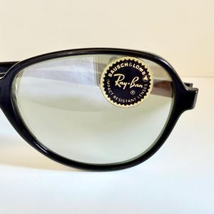 May include: Close-up of black-framed Ray-Ban sunglasses with green-tinted lenses. The Ray-Ban logo is visible on a circular sticker on the lens. The sunglasses have a classic, retro design.