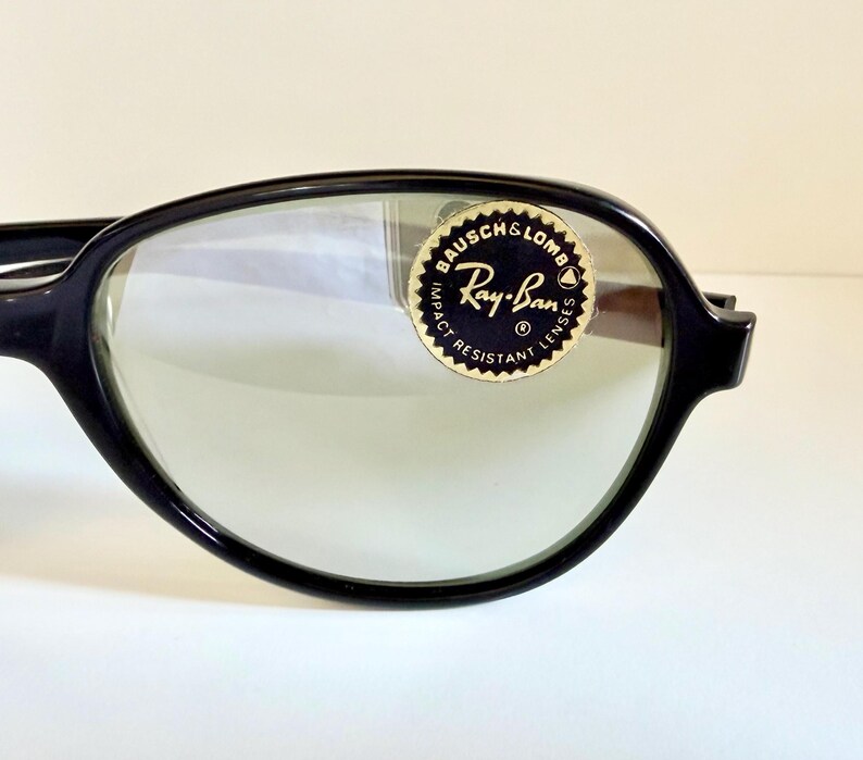 May include: Close-up of black-framed Ray-Ban sunglasses with green-tinted lenses. The Ray-Ban logo is visible on a circular sticker on the lens. The sunglasses have a classic, retro design.