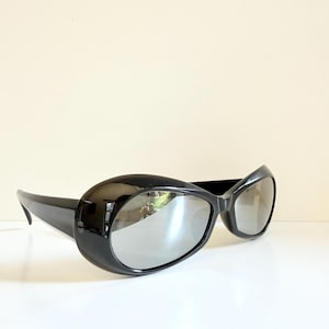 May include: Black sunglasses with oval frames and a glossy finish. The lenses have a mirrored appearance. The sunglasses are displayed against a neutral background, emphasizing their sleek design.