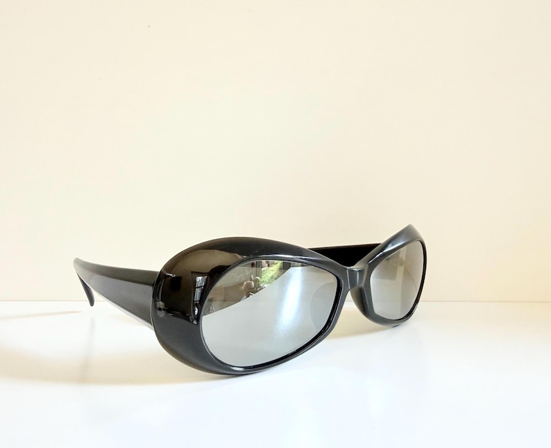 May include: Black sunglasses with oval frames and a glossy finish. The lenses have a mirrored appearance. The sunglasses are displayed against a neutral background, emphasizing their sleek design.