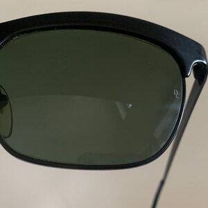 May include: Close-up of black sunglasses with dark green lenses. The sunglasses have a thick, curved top frame and thin metal arms. A small logo is visible on the right lens. The lenses are slightly reflective.
