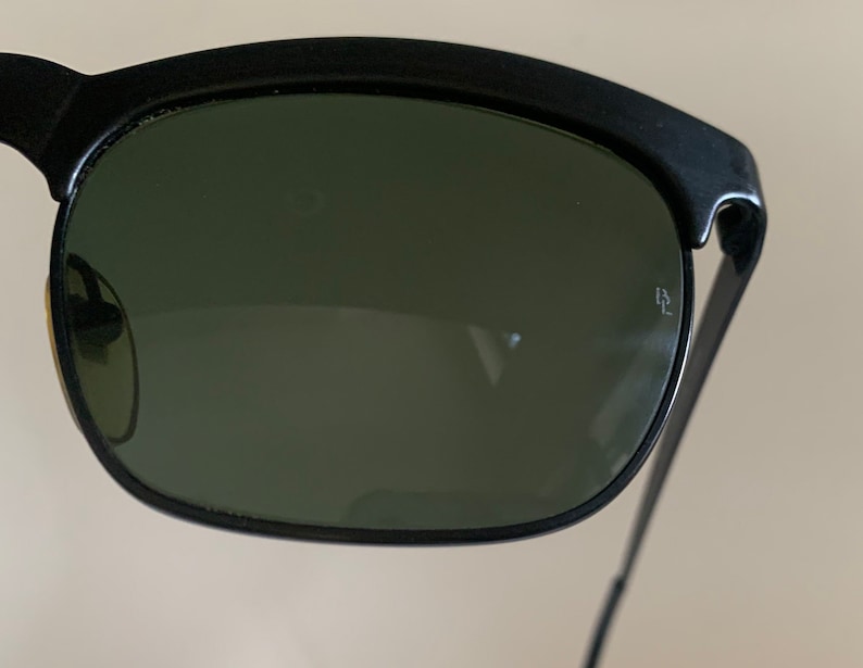 May include: Close-up of black sunglasses with dark green lenses. The sunglasses have a thick, curved top frame and thin metal arms. A small logo is visible on the right lens. The lenses are slightly reflective.